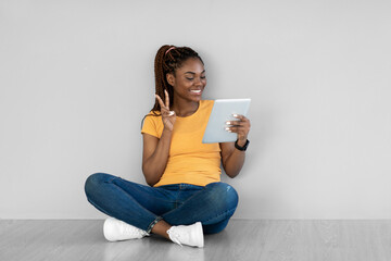 Online communication. Charming young black woman video chatting on tablet pc, showing peace gesture...