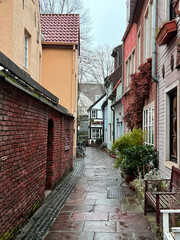 One of the narrow alleys in the Schnoor district in Bremen. At the left side a wall. On the other side the facades of houses. In front are benches and potted plants. It's a rainy day