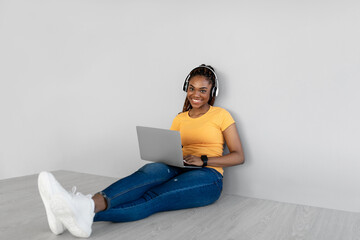 Full length of young black woman in headphones using laptop, sitting on floor against grey studio wall, free space