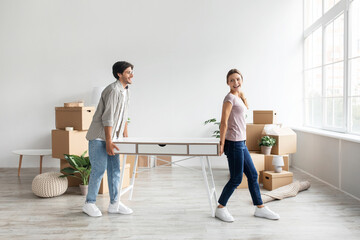 Cheerful millennial caucasian wife and husband carry table in room interior with cardboard boxes