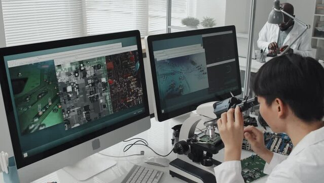 High angle shot of female electronics engineer using microscope and examining magnified images of circuit board on computers at lab desk
