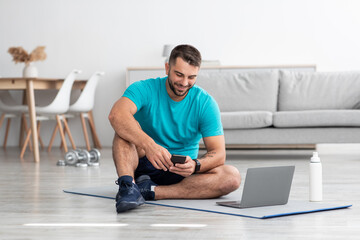 Cheerful millennial european man chatting on smartphone sits on mat in living room interior with bottle