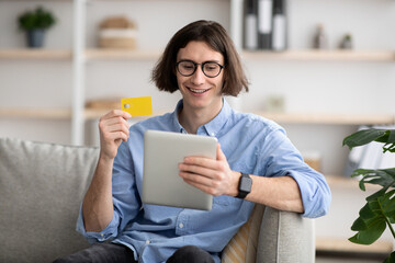 Online shopping concept. Happy young man holding debit credit card and using digital tablet, ordering food delivery