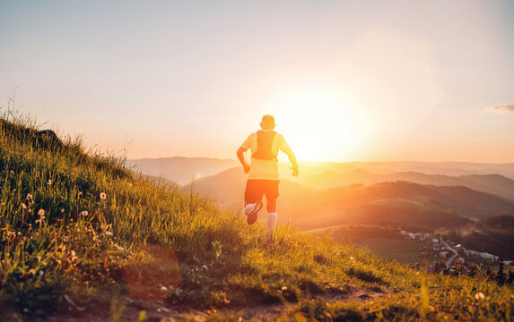 Active mountain trail runner dressed bright t-shirt with backpack running endurance marathon race by picturesque hills at sunset time back view photo. Sporty active people backlight concept image.