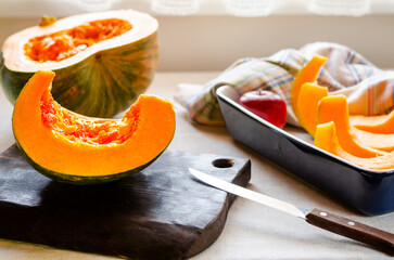 A bright orange pumpkin cut into pieces lies in a baking dish and on a chopping wooden board next...