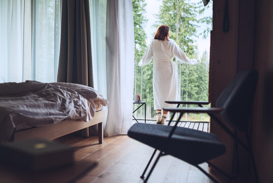 Woman dressed white bathrobe standing on forest house balcony and enjoying fresh air with nature view. Inside the Scandinavian interior design room with a not made bed. Living in wild concept image..