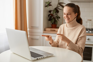 Portrait of attractive positive woman wearing beige sweater sitting in kitchen and working online on laptop, pointing at notebook display, expressing happiness.