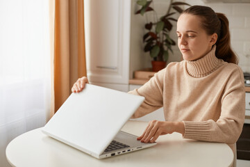 Indoor shot of young adult Caucasian woman wearing beige sweater sitting in kitchen and open laptop, being ready to start her online work, having concentrated expression.