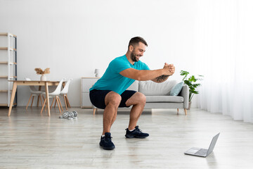 Fototapeta premium Happy millennial european guy doing leg exercises, squats and watching online lesson on pc