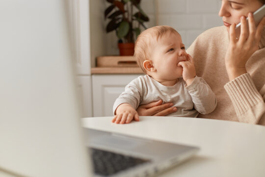 Cropped image woman wearing beige sweater sitting in kitchen and working online on laptop and talking via smartphone, looking after baby girl, calming down crying daughter.