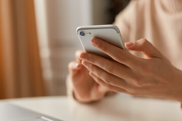 Cropped closeup portrait of woman wearing beige sweater sitting holding smart phone in hands, scrolling online on mobile phone, typing message.