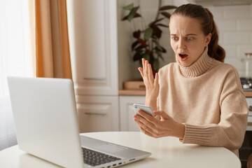 Portrait of shocked woman wearing beige sweater sitting in kitchen and working online on laptop and using smart phone, screaming, keeps mouth open, raised arm. - obrazy, fototapety, plakaty
