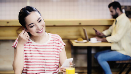 cheerful asian woman listening music near blurred man on background