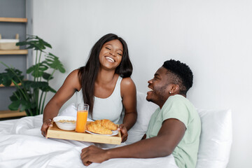 Obraz na płótnie Canvas Laughing happy millennial african american wife hold tray with breakfast for husband on bed in bedroom interior