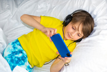 a happy smiling boy woke up and started playing a game on a smartphone while lying in bed, with headphones on his head. caucasian boy looking information video