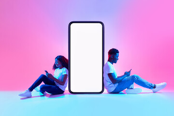Black couple sitting near big smartphone with mockup, using gadgets, advertising new app or website...