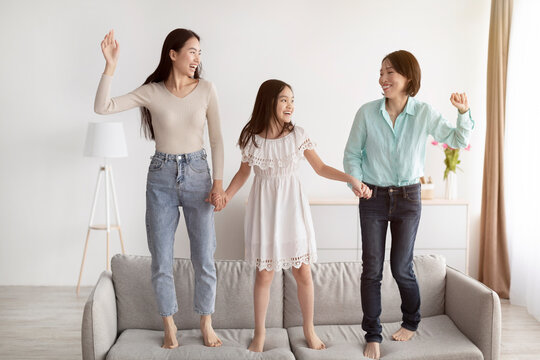 Happy Little Asian Girl With Her Mother And Granny Jumping On Couch, Having Fun In Living Room