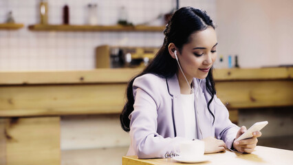 cheerful asian woman in suit listening music in earphones and using smartphone near cup of coffee in cafe