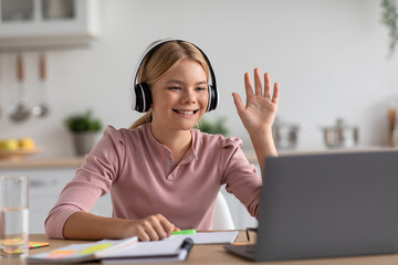 Happy caucasian teen girl pupil in headphones waving hand studying with laptop at table in kitchen interior