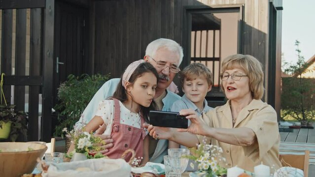 Waist-up of mature Caucasian woman holding smartphone, talking selfie with her little grandchildren and husband, sitting at table, talking and smiling outside summer house on sunny day