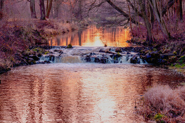 Wodospad na małej rzece płynącej przez las. Jest jesienny wieczór, światło zachodzącego słońca zabarwiło powierzchnię wody na czerwono.