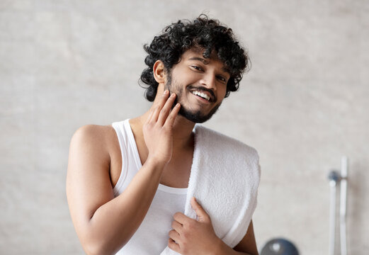 Portrait of happy indian man touching beard and smiling at camera, attractive young guy enjoying morning routine