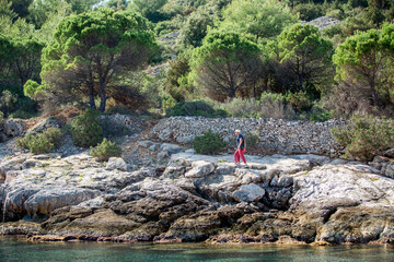 Mann mit Strohhut und roter Hose geht unter Bäumen über Felsen am Meer