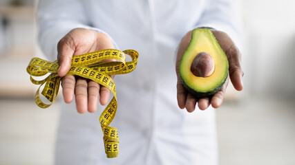Healthy lifestyle concept. Black female dietitian holding half of avocado fruit and measure tape,...