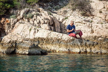 Digitaler Nomade mit Sonnenbrille und roter Hose sitzt auf Felsen am Meer und arbeitet am Laptop, Handzeichen und Emotionen Freude, Nahaufnahme