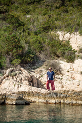 Digitaler Nomade mit Sonnenbrille, roter Hose und Laptop unterm Arm steht auf Felsen am Meer und blickt in die Ferne, Hochformat