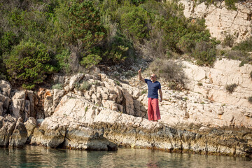 Digitaler Nomade mit Sonnenbrille, roter Hose und Laptop unterm Arm steht auf Felsen am Meer, Emotionen Freude & Gewinner