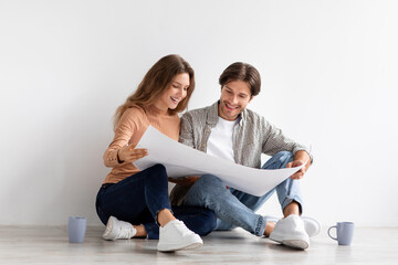 Fototapeta na wymiar Cheerful young european man and lady sitting on floor and looking at house plan, planning repairs and moving