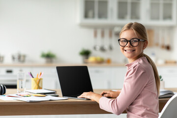 Fototapeta premium Cheerful caucasian teen schoolgirl in glasses look at camera at table with computer with blank screen