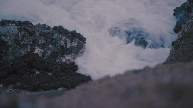 Small waves of white foamy water spill between the cracks of various barnicle, limpit covered rocks on a warm afternoon day