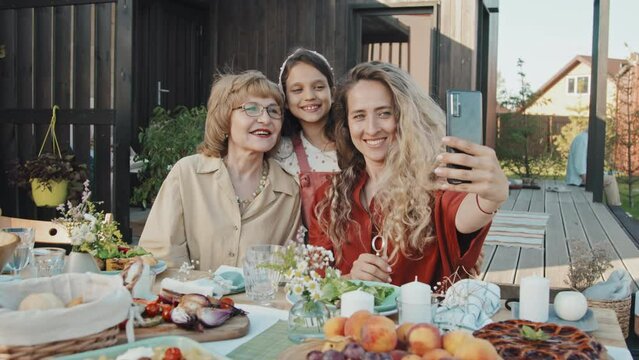 Medium of young Caucasian woman holding smartphone, talking selfie with her little daughter and mature mother, sitting at table outside summer house on sunny day