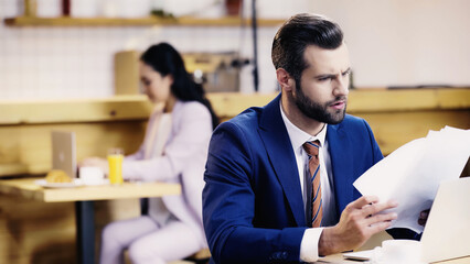 bearded businessman looking at documents near laptop and blurred woman in cafe