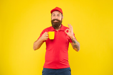 Happy bearded man in casual red cap and tshirt giving OK holding cup yellow background, coffee