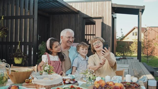 Waist-up of mature Caucasian woman holding smartphone, talking selfie with her little grandchildren and husband, sitting at table outside summer house on sunny day