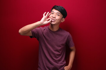 Portrait of asian man making delicious hand gesture, close up head and shoulder portrait against red background