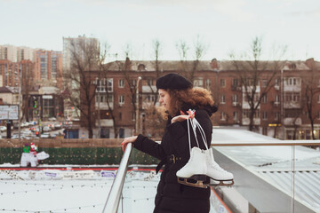Young curly-haired woman in a beret with ice skates looks down at the ice rink