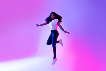 Fototapeta na wymiar Full length portrait of active energetic millennial African American woman jumping in neon light, copy space