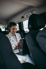 Good looking young business woman sitting on backseat in luxury car. She using her smart phone and smiling. Transportation in corporate business concept.