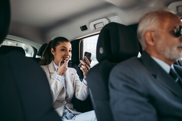 Good looking young business woman sitting on backseat in luxury car and fixing her makeup. Transportation in corporate business concept.