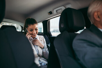 Good looking young business woman sitting on backseat in luxury car. She using her smart phone and smiling. Transportation in corporate business concept.