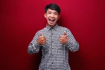 asian man wearing plaid shirt with happy expression showing his two thumbs up, on red background