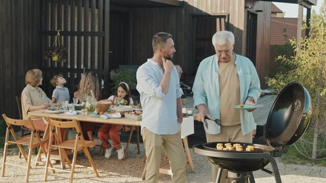 Medium long of senior Caucasian man and his adult son standing at grill in backyard of summer house, making BBQ and talking, women and children sitting at table on background
