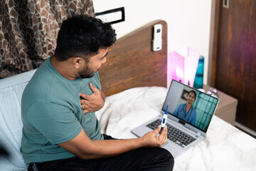 Indian sick man talking on video call by consulting doctor on laptop at bedroom - concept of...