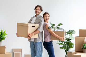 Happy millennial european woman and husband hold cardboard box with stuff and plants, stand back to...