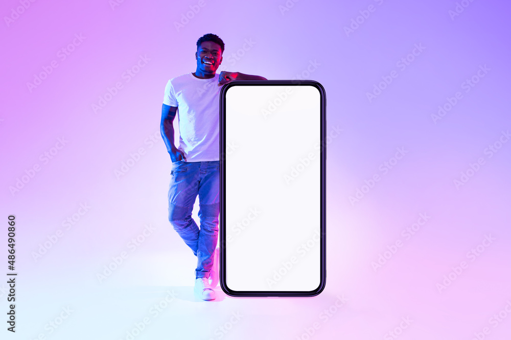 Poster smiling young black man standing near huge cellphone with empty white screen in neon light, mockup f