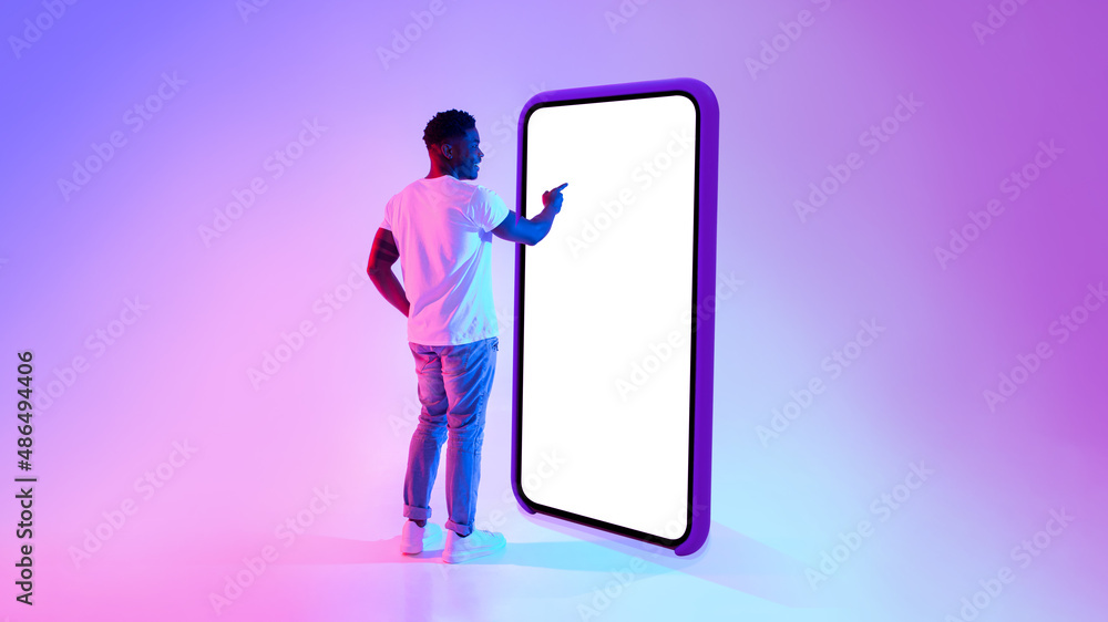 Poster Black guy standing near big cellphone with mockup for your app on screen, interacting with user interface in neon light